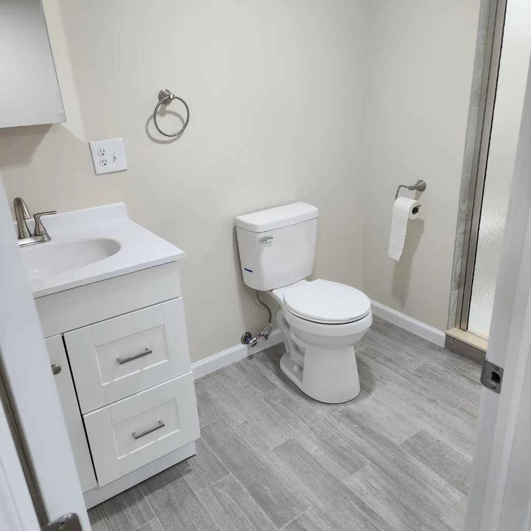 Compact bathroom with white vanity, toilet and wood-look porcelain tile floor by Link Services Pro in Worcester, MA