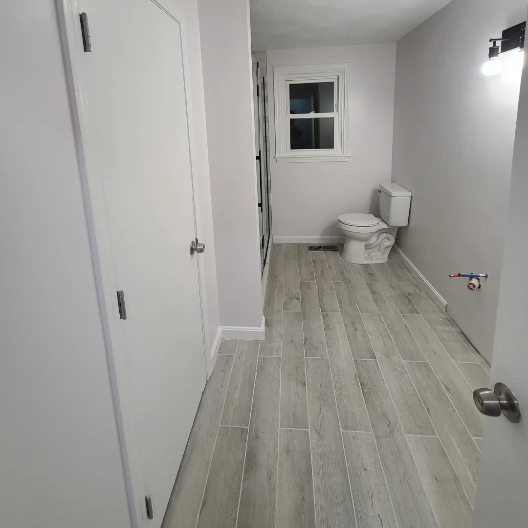 Wood-look porcelain tile flooring in bathroom with white toilet by Link Services Pro in Holden, MA